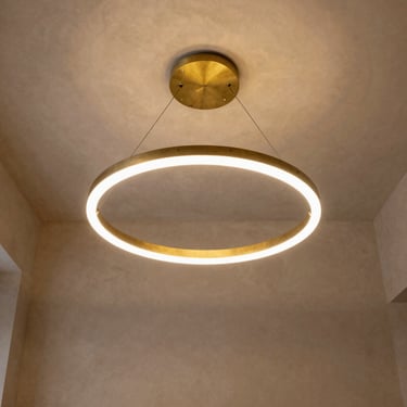 Minimalist hotel lobby with a single sculptural brass ring light suspended from a high plaster ceiling, warm golden glow, symmetric framing.