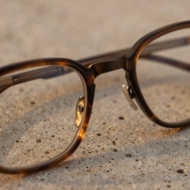 Extreme close-up of structural eyewear resting on porous travertine, warm low-intensity golden hour glow reflecting off bronze hinges.