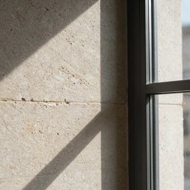 Close-up of textured limestone wall meeting dark metal window frame, high-contrast natural light, professional architectural detail.