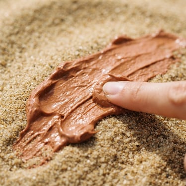Macro texture shot of thick terracotta-toned moisturizing cream being smeared across a warm sand surface, soft side-lit daylight