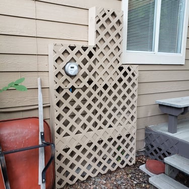 Tan garden lattice screen hiding outdoor utility meters on a house exterior next to a red wheelbarrow.