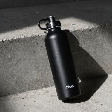 Crisp clinical shot of a black alkaline water bottle resting on raw concrete, sharp shadows, low-key lighting.
