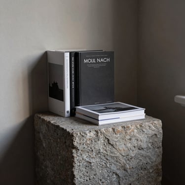 A quiet studio corner in Cheyenne, Wyoming, featuring a solid stone plinth holding linen-bound editorial books, soft indirect daylight filtering in.