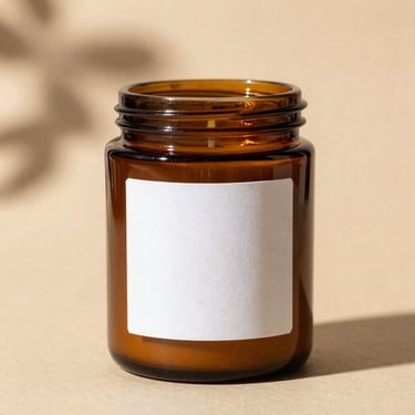 A minimal amber glass jar candle with a heavy paper label, soft botanical shadows on a warm background.