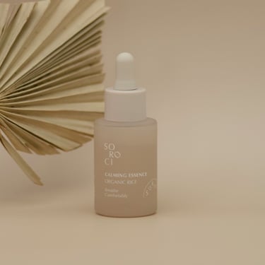 Sticlă de esență cu pipetă, prezentată lângă o frunză uscată decorativă, pe fundal bej uniform.