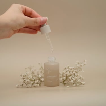 Esență calmantă cu pipetă, surprinsă în timp ce este deschisă, flancată de flori albe pe fundal bej.