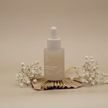 Sticluță de esență calmantă, cu pipetă albă, așezată central pe fundal bej, decorată cu flori albe delicate