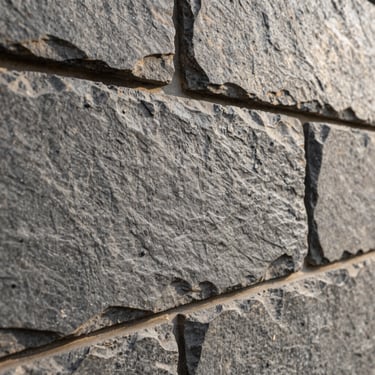 Macro detail shot of a textured grey stone wall, natural slate texture, sharp sunlight highlighting the relief, architectural detail