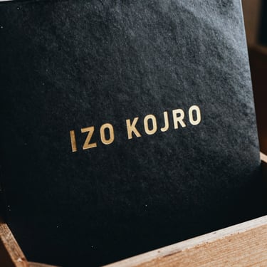 Tactile macro shot of a 12-inch vinyl record sleeve, matte black cardstock with debossed gold foil lettering, resting on a wooden crate.