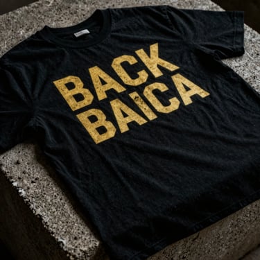 High-contrast photo of a screen-printed black cotton t-shirt with bold gold typography, draped over a concrete block under low-key lighting.