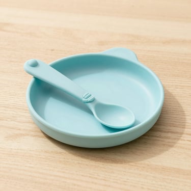A pastel-colored silicone baby plate and spoon on a clean oak table, soft side lighting.
