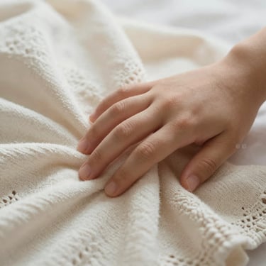 Close-up of a hand touching a soft organic cotton baby blanket with delicate knit details, warm natural light.