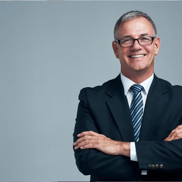 A smiling senior businessman wearing glasses and a suit standing with arms crossed against a grey background.