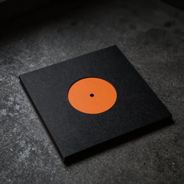 Minimalist vinyl packaging sleeve, matte black cardboard with a single debossed orange circle in the center, shot from a high angle on a dark concrete surface.