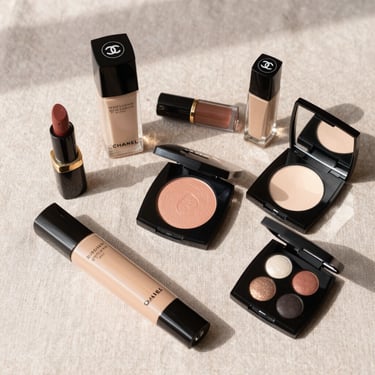 A beautifully arranged flatlay of luxury beauty products, Chanel and Westman Atelier, on a soft blush linen surface under soft morning light.