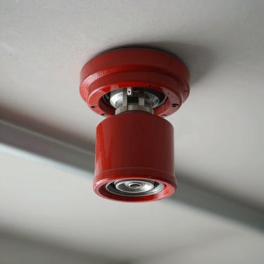 Close-up of a red fire sprinkler head mounted on a clean ceiling grid, surrounded by steel piping, high-contrast studio lighting, sharp focus.