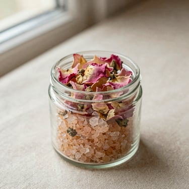 A glass jar of botanical bath salts with dried pink rose petals, linen texture background