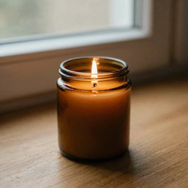 A hand-poured soy candle in an amber glass jar, lit, casting a warm soft glow on a wooden surface