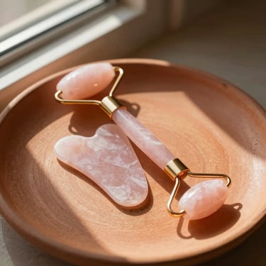 A rose quartz Gua Sha tool and facial roller resting on a textured terracotta plate, warm sunlight casting soft shadows