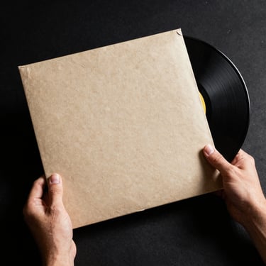 Album cover and vinyl record photographed together in controlled natural north-facing light, hands positioning the jacket at an angle, recycled paper texture and pressing detail visible, high-contrast studio setup with black background, flat-lay overhead framing.