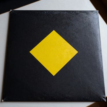 Close-up flat-lay of a vinyl record sleeve, deep black cover with a single electric-yellow geometric mark, studio table surface visible at edges, sharp overhead light