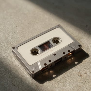 Tight close-up of a cassette tape resting on a concrete studio floor, warm tungsten sidelight casting a long shadow to the right, textured surface visible