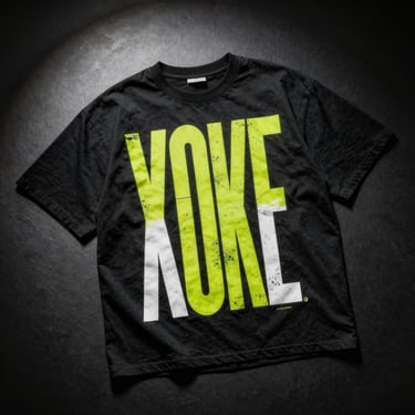 Flat lay of a black oversized tee on a matte dark surface, large bold graphic print across the chest in electric yellow-green and white, single overhead spot lighting with deep peripheral shadows, no model, no props