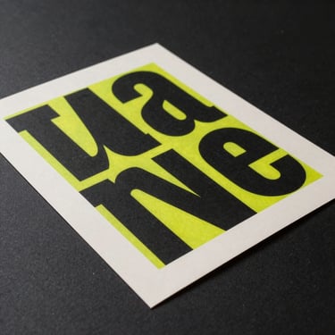 Tight product shot of a limited-edition art print lying flat on a dark matte surface, bold typographic artwork in black and electric yellow-green on off-white paper, slight curl at one corner revealing paper thickness, single focused overhead light, no frame shown