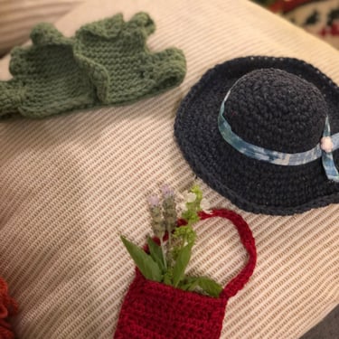 Piezas tejidas en crochet, chaleco verde, sombrero azul con cinta y bolsita roja con unas flores adentro sobre un cojin