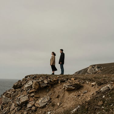 A couple standing quietly together on a windswept coastal cliff, muted earthy tones, overcast sky, cinematic wide-angle storytelling.