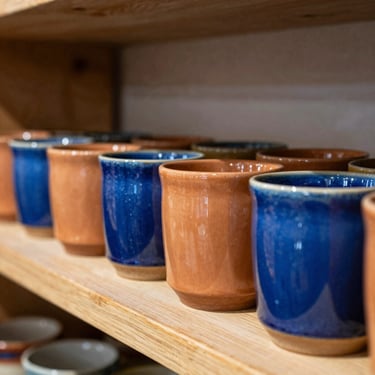 A row of heavy, freshly glazed stoneware mugs in terracotta and cobalt blue sitting on a dry wooden shelf