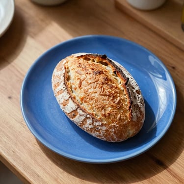 Overhead view of a messy kitchen table with a half-eaten sourdough loaf on a bright cobalt blue stoneware plate, sharp sunlight