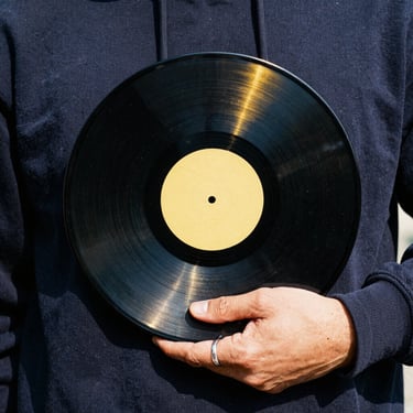 Close-up of a hand with a silver ring holding a vinyl record, gold and navy blue lighting, gritty fanzine aesthetic.