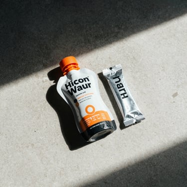 Overhead flat-lay on a pale concrete surface, a compact cycling nutrition gel flask with orange cap resting beside a folded foil packet, natural side-window light casting clean shadows, no props