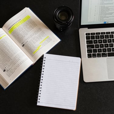 Top-down flat-lay of a neatly organized workspace, featuring an open textbook with yellow highlighting, a clean notepad, and a laptop displaying a structured document outline.