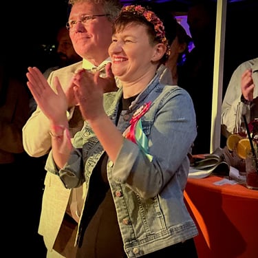 Smiling woman in a denim jacket and flower crown clapping at an outdoor evening event with friends.