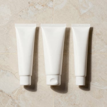 Flatlay of clean, white minimalist cosmetic packaging on a warm cream marble tile, soft diffused shadows.