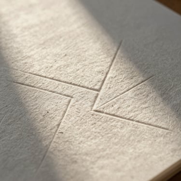 Extreme close-up of heavy cotton paper with blind-debossed geometric lines, warm morning light casting soft shadows across the textured surface.