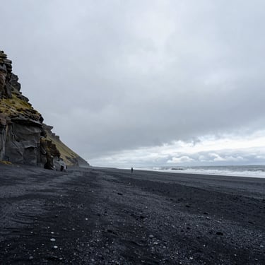 Ultra-wide environmental landscape of a rugged Icelandic volcanic black sand beach under heavy overcast sky, a tiny lone figure standing near the tide.