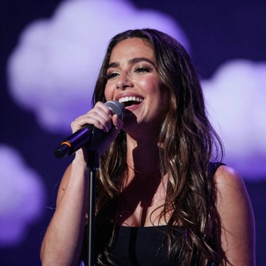 A portrait of a laughing woman singing into a microphone. She is surrounded by a soft bokeh of deep midnight indigo and pale cloud lavender lights. The style is candid and warm.