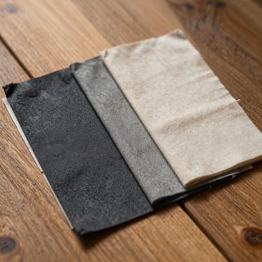 A close-up of physical paint swatches in warm charcoal and raw linen resting on a raw oak plank, natural morning light revealing subtle undertones.