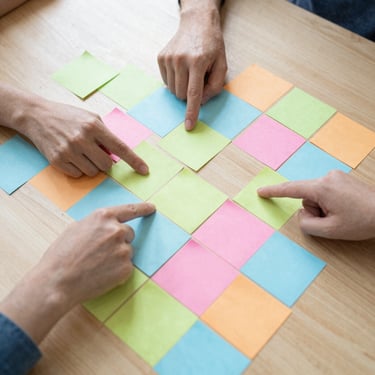 Overhead shot of a physical mapping session with colorful sticky notes on a wooden table, hands pointing to connections, bright daylight