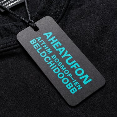 Macro shot of a custom neck tag on a heavy-gauge black cotton shirt, technical monospaced typography printed in electric teal