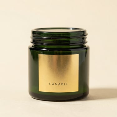 A close-up of premium cannabis packaging, a sleek dark green glass jar with a minimalist gold label, sitting on a clean warm cream surface, bright studio lighting