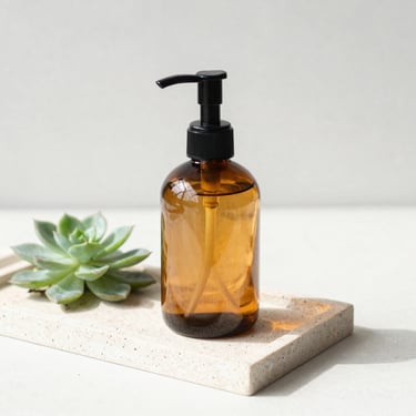 Aesthetic amber glass soap dispenser on a light stone tray next to a small green succulent, bright natural light