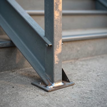 Detail of a steel stair stringer meeting a concrete landing — precise weld line visible, warm side light, minimal depth of field, no people or furniture