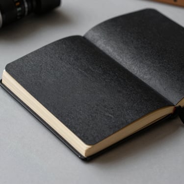 Stark close-up of a hardbound black linen notebook lying open on a grey desk, showing blank thick pages