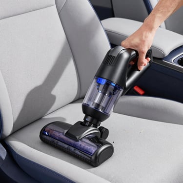 Medium close-up of a compact black handheld vacuum cleaner being used to clean crumbs from a minimalist grey fabric car seat, bright natural daylight, clean composition