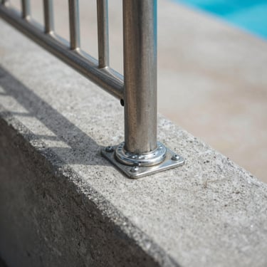 Close-up of a pool fence post base anchored into concrete pool coping, stainless steel anchor hardware visible, natural daylight, documentary framing, no people