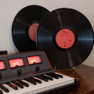 Moody studio interior with glowing red VU meters, a vintage synthesizer keyboard, and a dark vinyl record leaning against the wall.